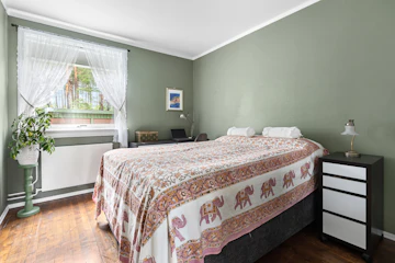 Bedroom with neatly made bed, window, lamp, plant, and desk-related furnishings.