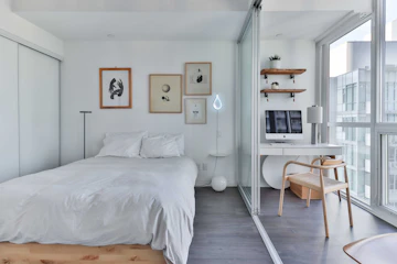 Apartment bedroom with bed, desk, window, lamp, and white interior.