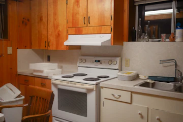 Simple kitchen with wooden cabinets, white appliances, sink, faucet, drawers, and stovetop.